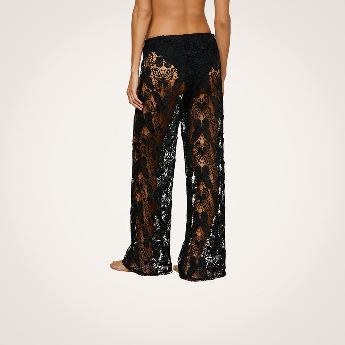 A person is shown from the back, barefoot and standing against a plain white background, wearing the Geraldine Cover-Up Pant—a pair of sheer black lace pants featuring a floral pattern that exudes a bohemian vibe.