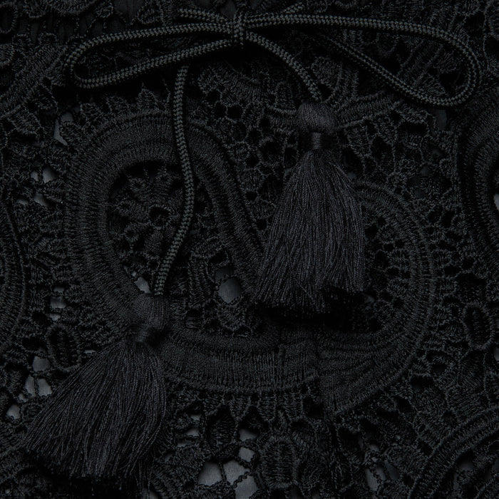 Close-up of the Geraldine Cover-Up Pant, featuring intricate black lace fabric adorned with decorative loops, a braided tie, and two tassels resting on top, creating a textured and elegant pattern reminiscent of resort wear.
