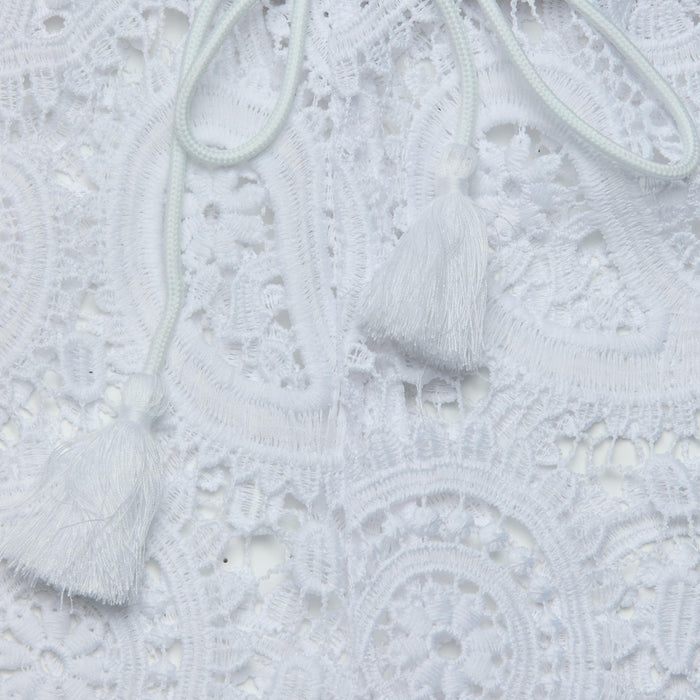 Close-up of the intricate white lace fabric of the Geraldine Cover-Up Pant, featuring two white tassels attached to thin cords resting on the lace—a perfect detail for resort wear or bohemian style.