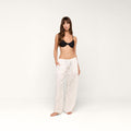 A woman stands against a plain white background, barefoot with her hands in the pockets of the Geraldine Eyelet Cover-Up Pant—a loose cotton eyelet pant with a tie waist—worn with a black bra, looking at the camera.