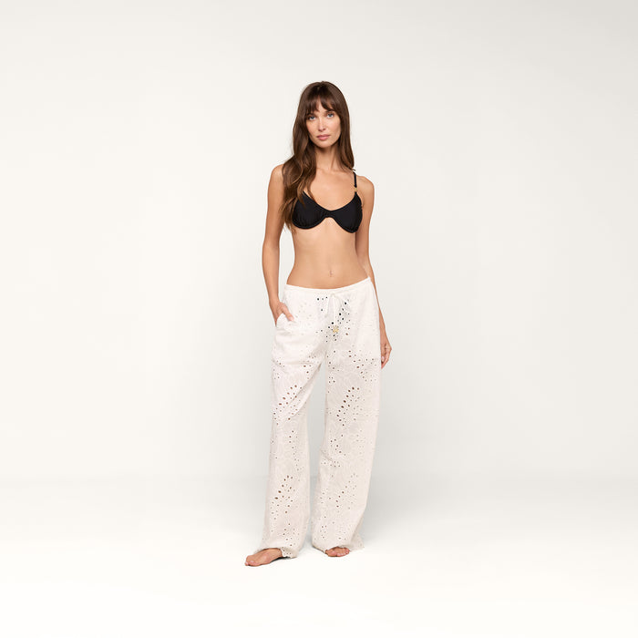 A woman stands against a plain white background, barefoot with her hands in the pockets of the Geraldine Eyelet Cover-Up Pant—a loose cotton eyelet pant with a tie waist—worn with a black bra, looking at the camera.