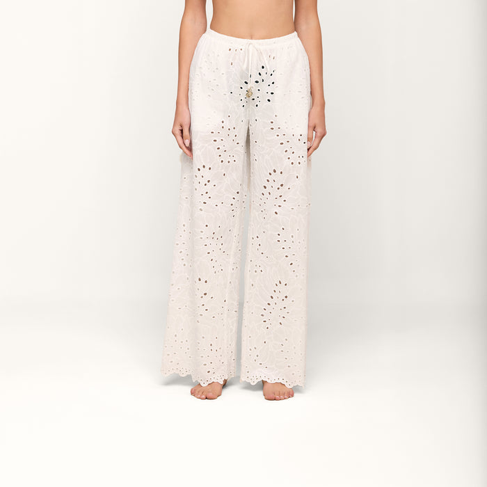 A person stands barefoot against a plain white background, wearing the Geraldine Eyelet Cover-Up Pant—a loose, white cotton style featuring an eyelet lace design, scalloped hem, and tie waist. The upper body and head are not visible.