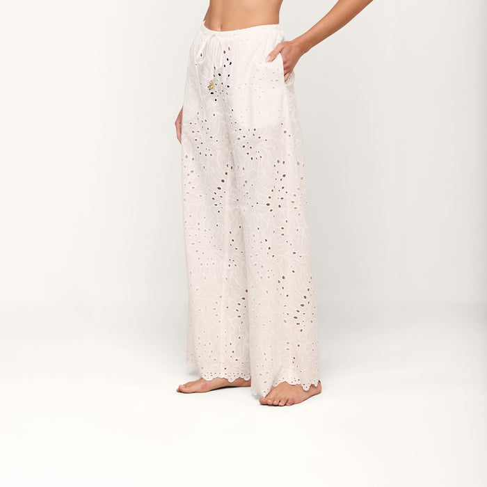 Barefoot, a person models the Geraldine Eyelet Cover-Up Pant—high-waisted, wide-leg cotton pants featuring a tie waist. The image focuses on the lower torso and legs set against a plain, light background.