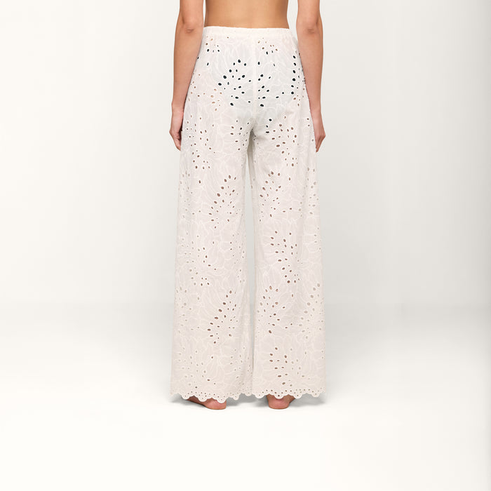 A person stands barefoot, facing away, wearing the Geraldine Eyelet Cover-Up Pant—high-waisted, white cotton eyelet pants featuring a scalloped hem. Elegant and designed for resort wear, these semi-sheer pants showcase a delicate floral cutout pattern.