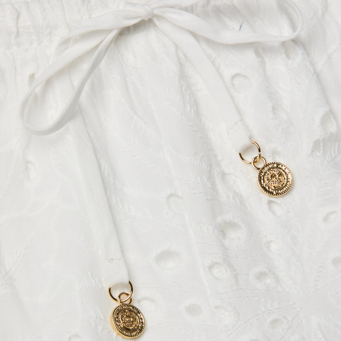 Close-up of the Geraldine Eyelet Cover-Up Pant, crafted from white cotton eyelet fabric with a tie waist and drawstring bow. Two gold decorative pendants accent the drawstring ends, bringing an elegant touch to your resort wear ensemble.