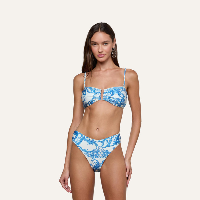 A woman wearing the Gianna Brazilian Bikini Bottom, featuring a blue baroque print with high-waisted and cheeky cut, stands against a plain light background.