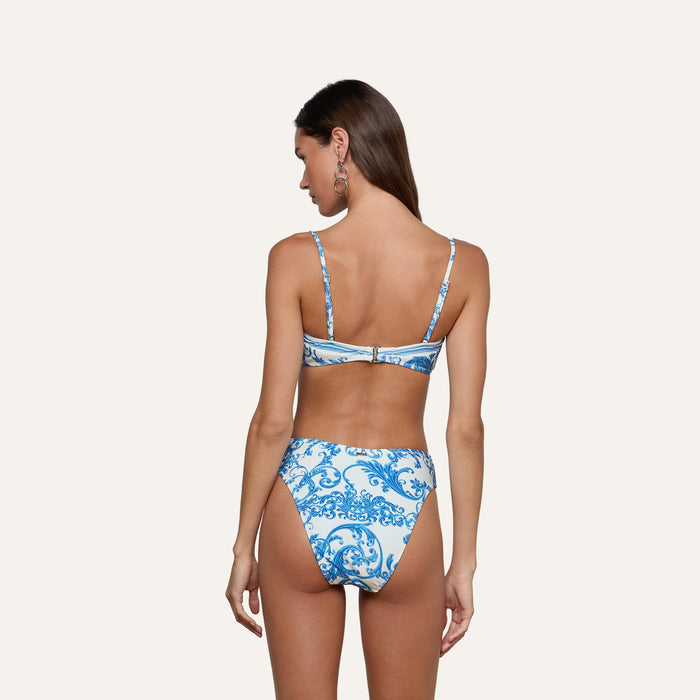 A woman with long brown hair stands against a plain light background, facing away and wearing the Gianna Brazilian Bikini Bottom—a blue and white bikini featuring an ornate swirl pattern and a cheeky cut.