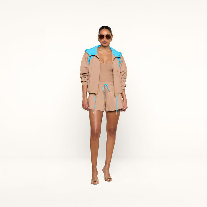 A woman stands confidently facing forward against a plain white background, wearing the Grayson Nylon Short paired with a tan crepe nylon jacket featuring a blue hood and a matching tan top. She accessorizes her look with sunglasses and clear heels.
