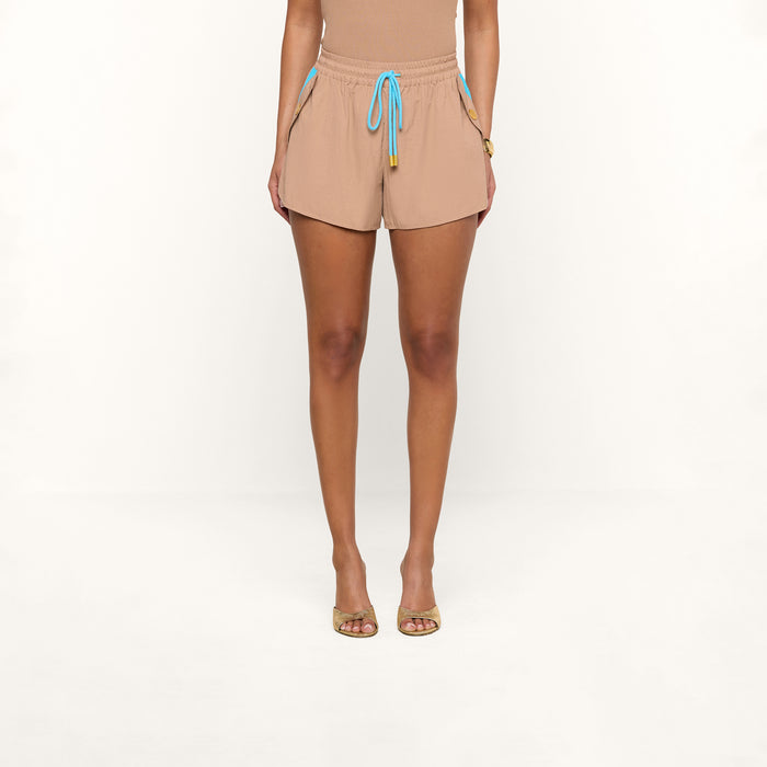 A person stands facing forward, wearing the Grayson Nylon Short in beige—crafted from water-repellent crepe nylon and featuring blue drawstrings—paired with a matching beige top. They wear open-toed sandals, set against a plain white background.