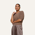 A woman with medium skin tone stands against a plain background, wearing the Guinevere Cropped Pullover—an ultra soft knit brown crewneck sweater—paired with tan satin trousers. She accessorizes with gold hoop earrings and has her hair pulled back, her right arm bent with her hand near her neck.