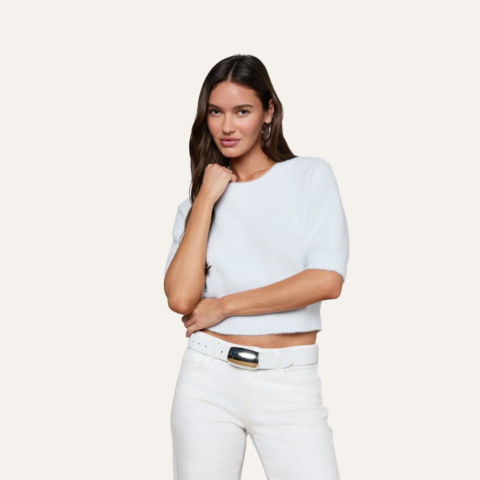 A woman with long brown hair wears the Guinevere Cropped Pullover—a light blue soft knit crewneck sweater with a cropped silhouette—paired with white pants, posing with one hand touching her neck against a plain light background.