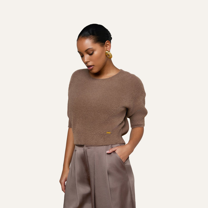 Wearing the Guinevere Cropped Pullover—a brown, ultra-soft crewneck sweater with a cropped silhouette—a woman stands against a plain light background, one hand in her pocket and her gaze cast downward, paired with high-waisted trousers.