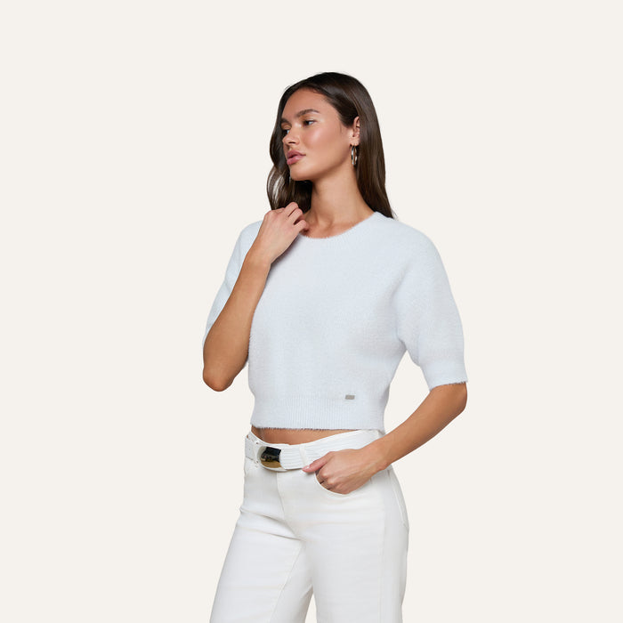 A woman wearing the Guinevere Cropped Pullover and white pants stands against an off-white background, resting one hand on her neck and the other in her pocket.