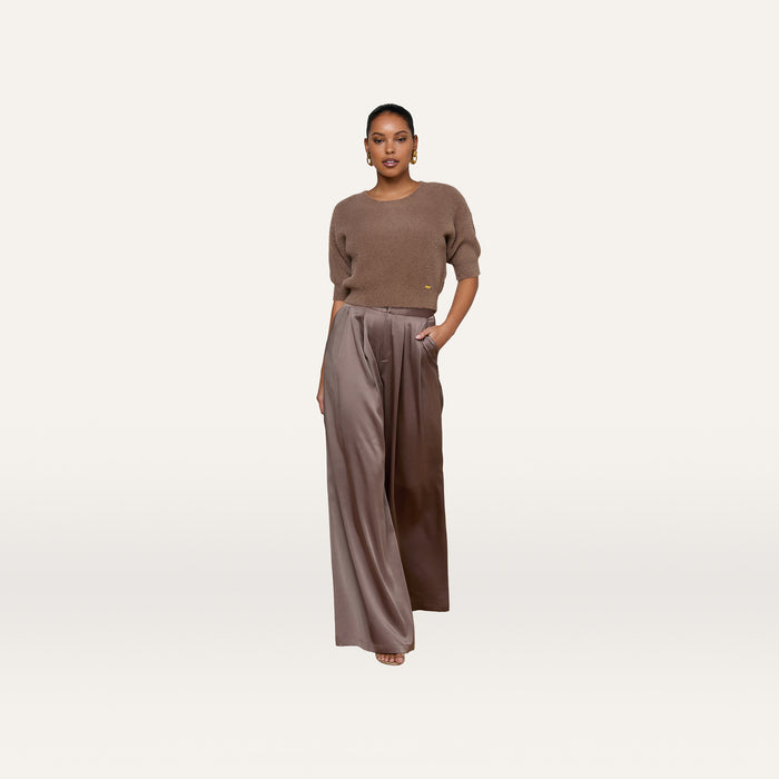 A person stands facing forward against a plain white background, wearing the Guinevere Cropped Pullover—an ultra soft knit brown short-sleeve top—paired with wide-legged, high-waisted brown satin pants with a cropped silhouette, one hand in their pocket.