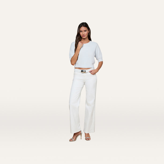 A woman stands confidently against a plain white background wearing the Guinevere Cropped Pullover, a light blue soft knit crewneck sweater with a cropped silhouette, paired with white wide-leg pants and heeled sandals.