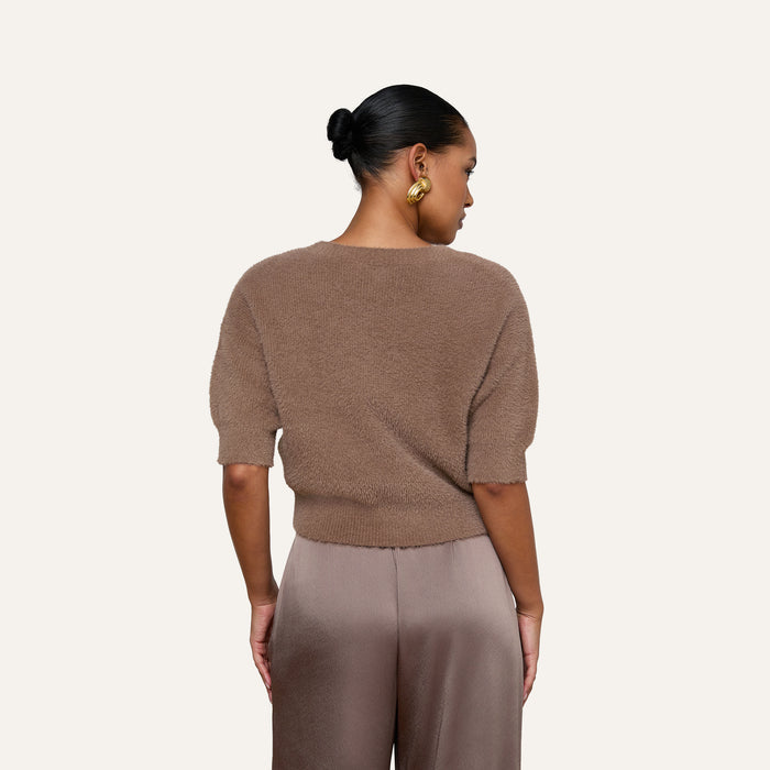 A woman with her hair in a bun and gold hoop earrings is turned away from the camera, revealing the back of the Guinevere Cropped Pullover—a brown ultra-soft knit short-sleeve crewneck sweater with a cropped silhouette—paired with taupe satin pants, set against a plain light background.
