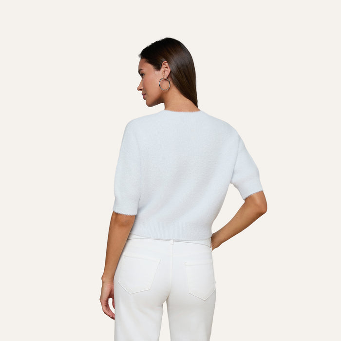 A woman stands with her back to the camera, wearing the Guinevere Cropped Pullover—a white soft knit crewneck sweater—paired with white jeans. She has straight brown hair, hoop earrings, and one hand resting on her hip against a plain, light-colored background.