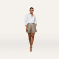 A woman stands against a plain background, wearing a white button-up shirt, the Hadleigh Linen-Blend Short in tan, and beige heeled sandals. She has one hand in her pocket and a relaxed expression.