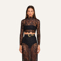 A woman wearing the Henriette Cropped Lace Top—a sheer, black Chantilly lace long-sleeved design—paired with high-waisted black shorts featuring a wide belt with a bold gold buckle. The outfit is distinguished by its intricate lace detailing and scalloped hems.