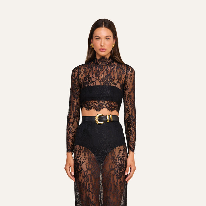 A woman wearing the Henriette Cropped Lace Top—a sheer, black Chantilly lace long-sleeved design—paired with high-waisted black shorts featuring a wide belt with a bold gold buckle. The outfit is distinguished by its intricate lace detailing and scalloped hems.