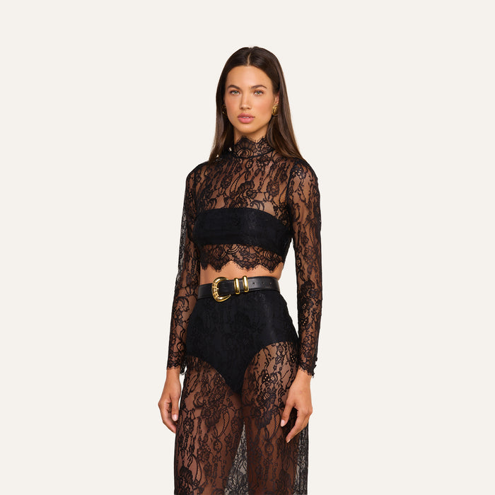 Wearing the Henriette Cropped Lace Top, a woman stands against a plain light background. She pairs the sheer black Chantilly lace long-sleeved top with a black bandeau, high-waisted black underwear, and finishes the look with a black belt featuring a large gold buckle.