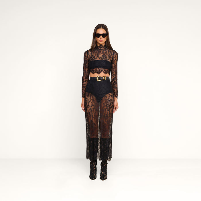 Woman stands against a plain white background wearing the sheer black Henriette Cropped Lace Top with scalloped hems, high-waisted black shorts, a black bandeau, black boots, a wide belt with a gold buckle, and dark sunglasses.