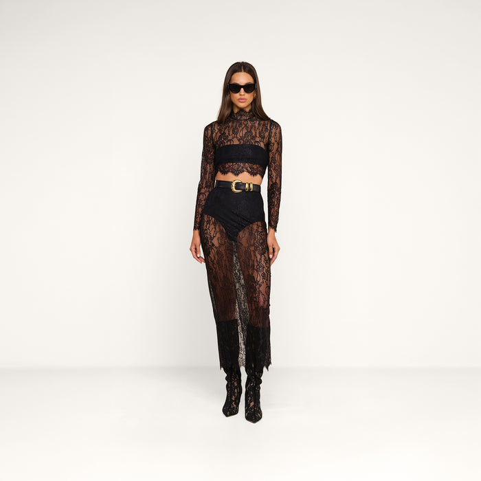 A woman in a black Henriette Cropped Lace Top with long sleeves, scalloped hems, a sheer skirt, high-waisted shorts, a gold-buckled belt, heeled boots, and sunglasses stands against a plain white background.