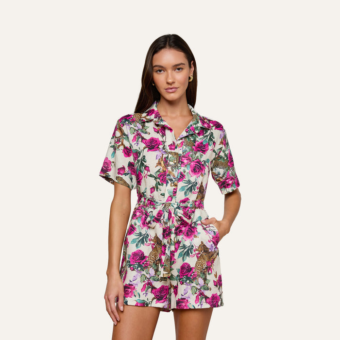 A woman stands with her hand in her pocket, facing forward against a plain light background, wearing the Hestia Cover-Up Romper—a short-sleeved, button-up linen romper featuring a pink floral and leopard print pattern.