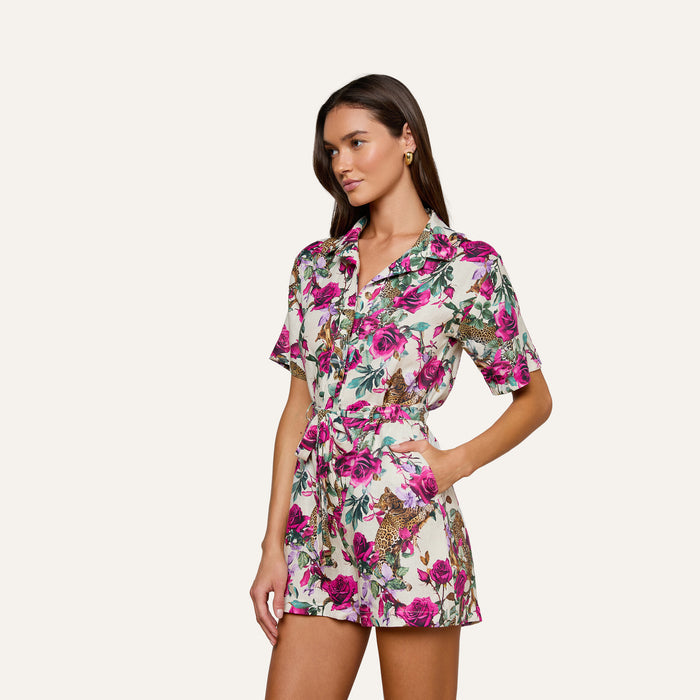 A woman poses with one hand in her pocket against a light background, wearing the Hestia Cover-Up Romper—a short-sleeve, button-up piece featuring a vibrant floral and rose leopard print.