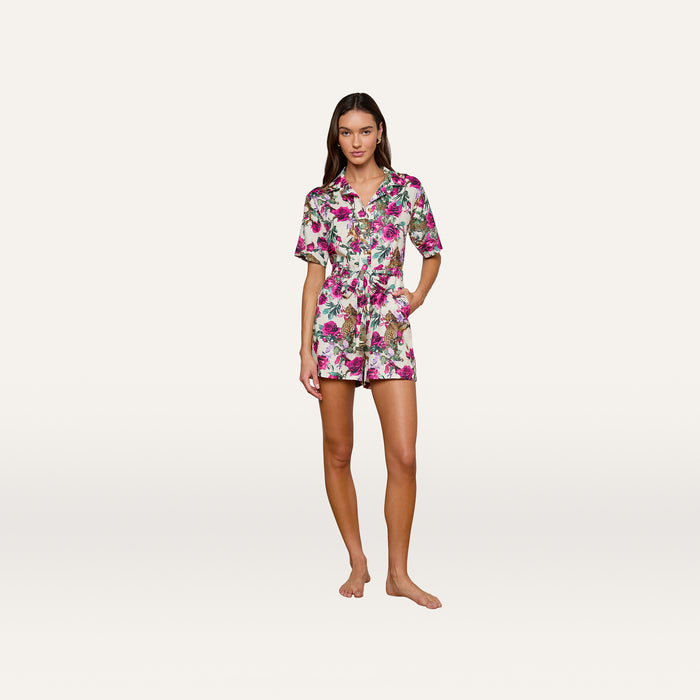 A woman stands barefoot against a plain white background, hands in her pockets, wearing the Hestia Cover-Up Romper—a short-sleeved linen piece featuring a pink and green floral pattern.