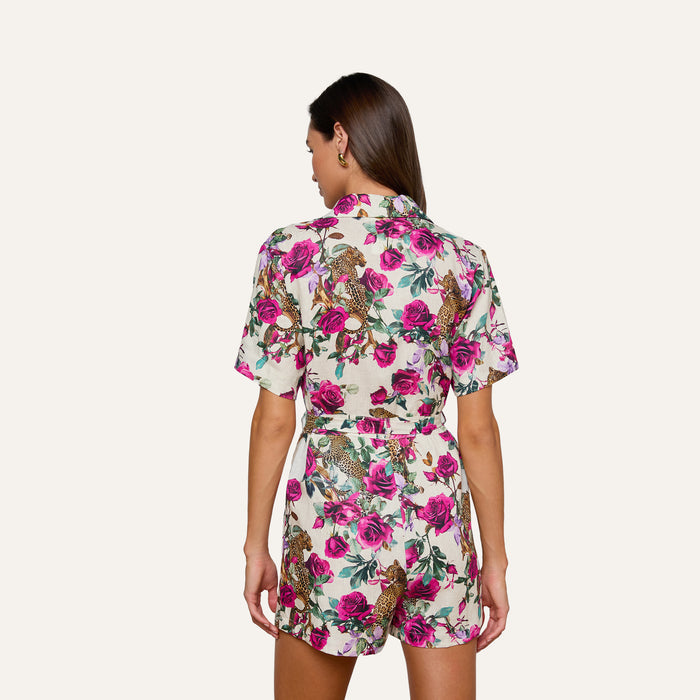 A woman faces away, wearing the Hestia Cover-Up Romper—a short-sleeved linen piece featuring a vibrant floral and rose leopard print in shades of pink, green, and brown on a white background. Her brown hair is worn down as she stands against a plain light backdrop.