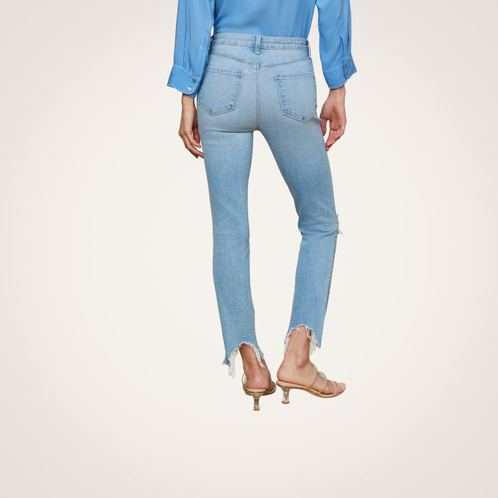 A woman stands with her back to the camera against a plain white background, wearing the High Line Distressed Skinny Jean in a light blue vintage wash with frayed hems, paired with a blue long-sleeve shirt and beige heeled sandals.