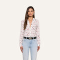 A woman with long brown hair and a neutral expression stands against a plain background, wearing the Holly Blouse—a slim fit blouse featuring a lipstick print design—paired with light blue jeans and a black belt.