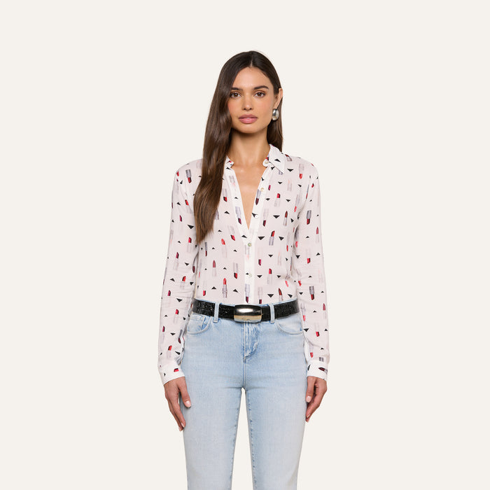 A woman with long brown hair and a neutral expression stands against a plain background, wearing the Holly Blouse—a slim fit blouse featuring a lipstick print design—paired with light blue jeans and a black belt.