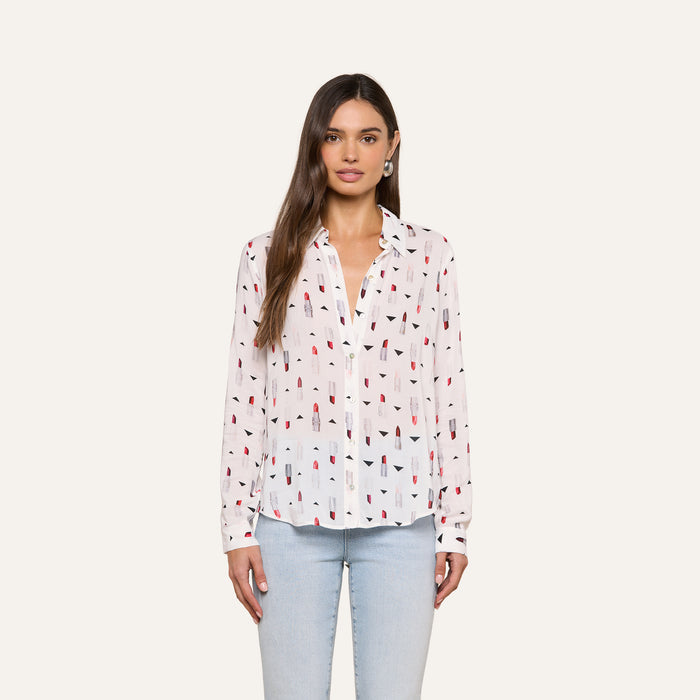 A woman with long brown hair wears the Holly Blouse—a slim fit shirt featuring a playful lipstick print, long sleeves, and a button-down front—paired with light blue jeans, standing against a plain light background.