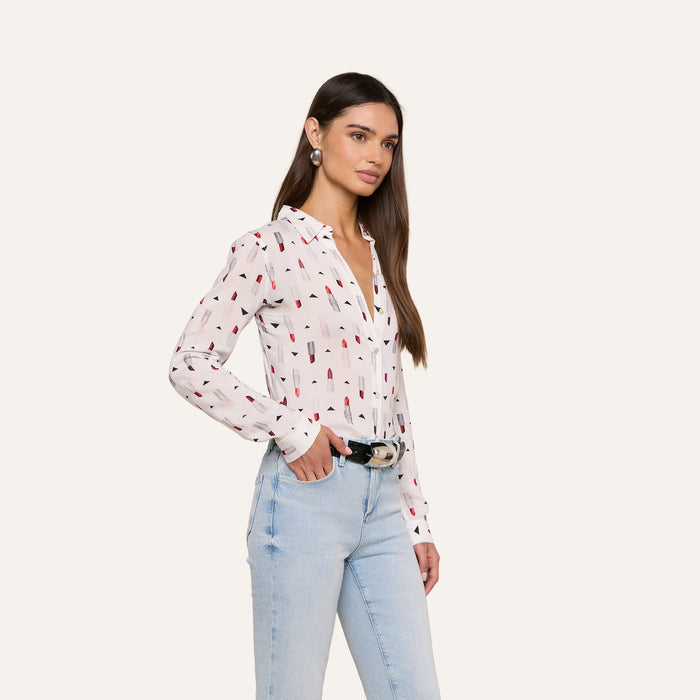 A woman with long brown hair wears the slim-fit Holly Blouse, featuring a lipstick print, paired with light blue jeans and a black belt. She stands with one hand in her pocket against an off-white background.