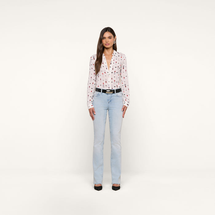 A woman stands against a plain white background, wearing the Holly Blouse—a slim fit top featuring a red and black pattern—paired with light blue jeans, a black belt, and black shoes. She has long, straight brown hair and her hands rest by her sides.
