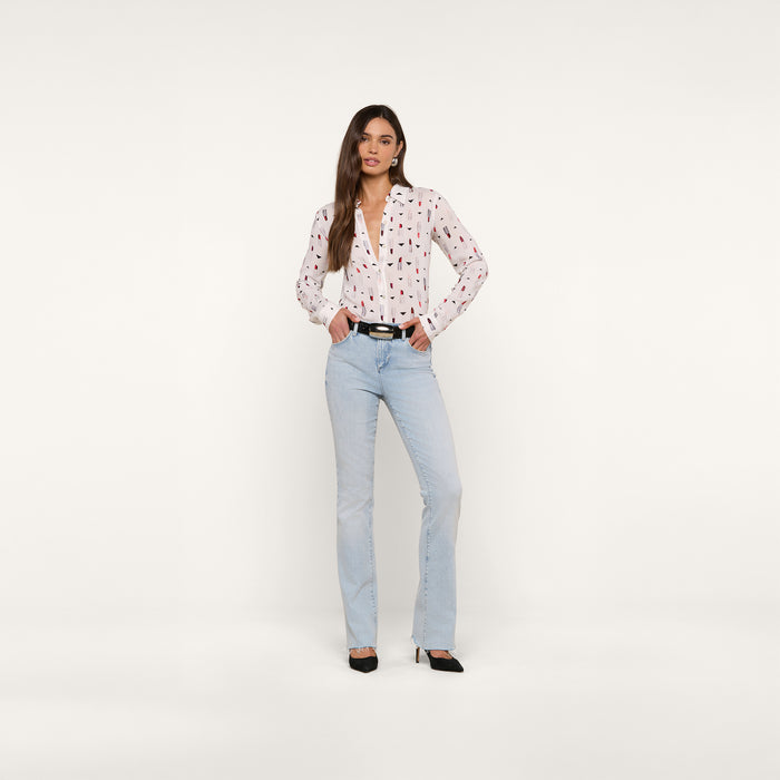 A woman stands against a plain white background, wearing the Holly Blouse paired with light blue jeans, a black belt, and black heels. She has long brown hair and strikes a relaxed pose with one hand in her pocket.