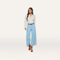 A woman is wearing a white long-sleeve blouse, the Houston Cropped Wide-Leg Jean in light blue stretch denim, a brown belt, and brown heeled sandals, standing against a plain white background.