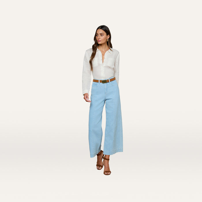 A woman is wearing a white long-sleeve blouse, the Houston Cropped Wide-Leg Jean in light blue stretch denim, a brown belt, and brown heeled sandals, standing against a plain white background.