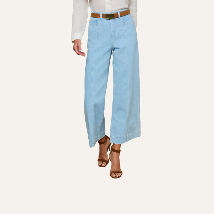 A person wearing the Houston Cropped Wide-Leg Jean in light blue stretch denim, styled with a brown belt, a white shirt, and brown sandals, stands against a plain background.