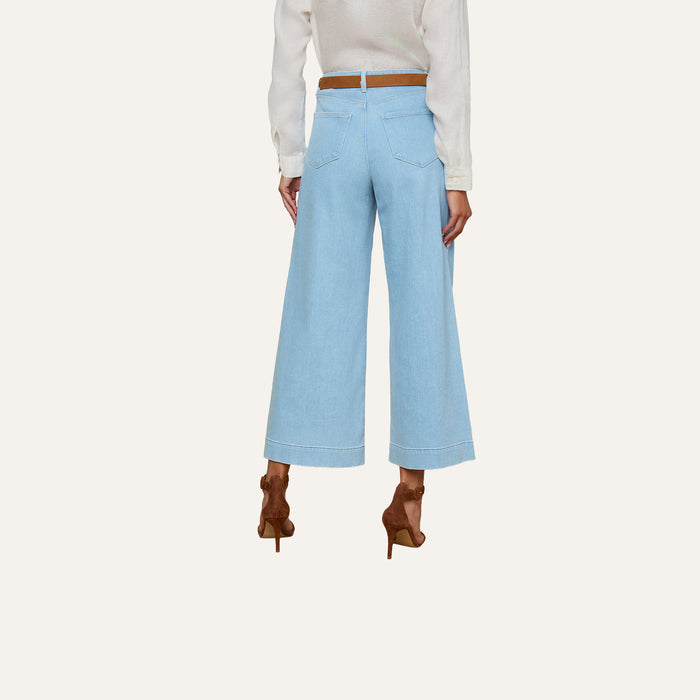 A woman is shown from the back against a plain background, wearing the Houston Cropped Wide-Leg Jean in light blue stretch denim, styled with a white long-sleeve top, a brown belt, and brown high-heeled shoes.