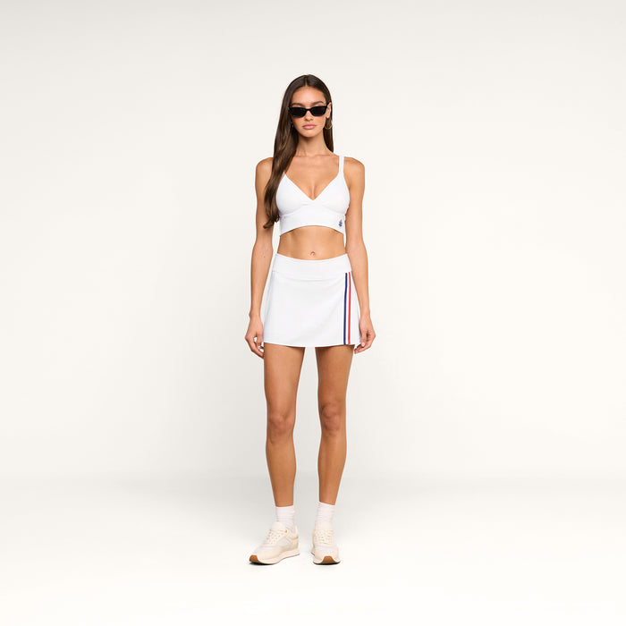 A woman stands against a plain white background wearing a white sports bra, the Iga Tennis Skort in white performance fabric with side stripes, white sneakers, and sunglasses.
