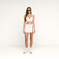 A woman stands against a plain white background wearing a white sports bra, the Iga Tennis Skort in white performance fabric with side stripes, white sneakers, and sunglasses.