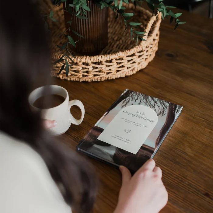 The back of a woman looking at In the Grips of Grace study cover on a brown wooden table with a coffee cup next to it and a woven basket with a potted plant in it
