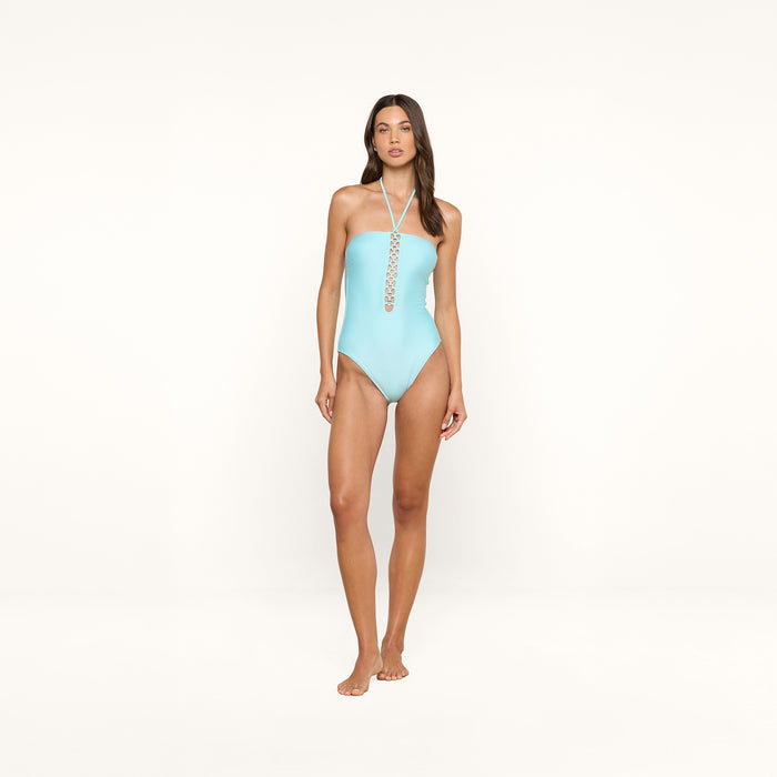 A woman stands against a plain white background, wearing the Indore One-Piece Swimsuit in ice blue with a lace-up front detail.