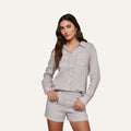 Wearing the Isla Linen Shirt in light grey, a woman with one hand in her pocket stands against a plain background; the button-down shirt features long sleeves and is paired with matching shorts.