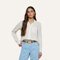 A woman with long brown hair wears the Isla Linen Shirt, a white button-down, paired with light blue high-waisted jeans as she stands with one hand on her hip against a plain background.