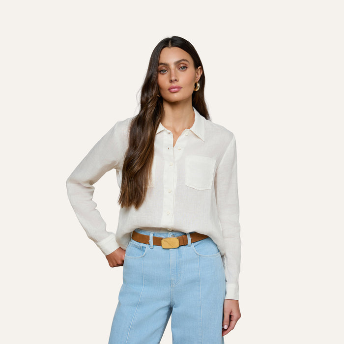 A woman with long brown hair wears the Isla Linen Shirt, a white button-down, paired with light blue high-waisted jeans as she stands with one hand on her hip against a plain background.