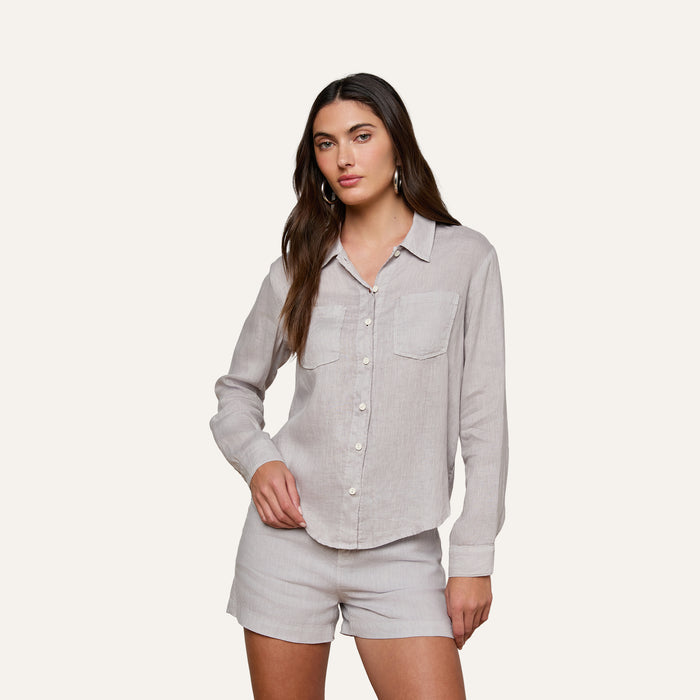 A woman stands against a plain background wearing the Isla Linen Shirt, a light grey, long-sleeve button-down with two front pockets, paired with matching shorts.
